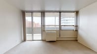 This one-bedroom, one-bathroom unit in Ann Arbor, Michigan offers a well-organized single-floor layout with a clean, functional design throughout. The living area is generously proportioned, finished with light-toned carpeting and bright white walls that create an open, airy atmosphere. A wall of floor-to-ceiling sliding glass doors dominates one side of the living room, providing direct access to a private balcony and framing views of the surrounding Ann Arbor cityscape, including neighboring buildings and open sky. A baseboard heating unit and a wall-mounted air conditioner ensure year-round climate control.

Adjacent to the living area, the kitchen features light-colored cabinetry extending to the upper walls, offering substantial storage. White appliances — including a refrigerator, electric range, and oven — are paired with a single-basin sink and continuous countertop workspace. The kitchen flooring transitions to a hard surface, distinguishing it from the carpeted living space.

A built-in shelving unit near the kitchen provides additional storage and organizational convenience. The bathroom includes a tiled tub-and-shower combination equipped with safety grab bars, a wood-framed vanity mirror, an oak vanity cabinet with drawers, and standard fixtures — all finished in neutral, easy-to-maintain tones throughout.