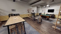 This single-floor recreational lounge offers a well-appointed social space designed for entertainment and relaxation. The room features an industrial-inspired aesthetic, highlighted by an exposed ceiling with visible ductwork and pendant lighting that casts a warm, inviting glow throughout the space.

At the heart of the room is a full-sized pool table with a teal felt surface, complemented by a cue rack mounted nearby. A tall bar-height wooden table with matching stools anchors one side of the room, providing a casual spot for games and conversation. Open wooden shelving units display books and decorative items, adding both function and character to the space.

A cozy seating area features modern lounge chairs paired with a small side table and a draped throw blanket, creating a comfortable retreat within the larger room. Patterned area rugs in a chevron design define different zones across the dark hardwood-style flooring, adding visual warmth and structure.

The walls are finished with a two-tone treatment — crisp white above and a muted yellow-gold below — and are adorned with framed artwork and prints that contribute to the space's lively, curated atmosphere. Additional furniture, including a dark console table and a beverage display unit, round out the room's amenities, making this lounge a versatile and engaging communal destination.