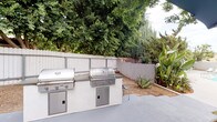 Located at 8836 Highland Ave in Whittier, California, this single-level outdoor amenity space is thoughtfully designed for al fresco entertaining and relaxation. The centerpiece of the space is a substantial built-in outdoor kitchen station, equipped with multiple stainless steel grills and storage compartments set into a clean, plastered counter base — ideal for hosting gatherings of any size. The expansive concrete patio provides ample room for movement and socializing, while a dedicated dining area features a wooden table, white chairs, and a large navy blue shade umbrella, creating a comfortable spot for outdoor meals. Lush, mature trees line the perimeter fence, offering natural privacy and shade, while tropical plantings including banana trees and agave add vibrant, layered greenery throughout. A secondary grill station is positioned adjacent to the dining area, further enhancing the cooking and entertaining capabilities of the space. A swimming pool is visible nearby, seamlessly connecting the outdoor kitchen and lounge areas to a full recreational amenity. The space is enclosed by a combination of white-painted wood fencing and tiled block walls with steel railings, providing both security and an open, airy feel suited to the Southern California climate.
