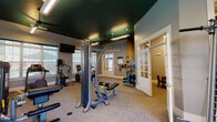 This fitness center is designed to support a comprehensive workout experience within a single-level layout. The main exercise area is distinguished by its bold dark-green ceiling, grey walls, and neutral carpet flooring, creating a polished yet energetic atmosphere. Recessed lighting and suspended linear fixtures, complemented by ceiling fans, ensure the space is well-lit and ventilated throughout.

The cardio zone is lined with large windows overlooking an outdoor pool area, providing natural light and views while accommodating treadmills, elliptical trainers, and a recumbent bike. Wall-mounted flat-screen televisions are positioned throughout for entertainment during workouts. The strength training section features a multi-function cable machine, a weight bench, and multiple dumbbell racks with a wall-length mirror enhancing the sense of space. A dedicated rack holds medicine balls, stability balls, and resistance equipment for functional training.

A separate adjoining room, accessible through glass-paned French doors, serves as a lounge or activity area with small tables, chairs, and playful wall art, offering a casual retreat. The overall design balances functionality with a clean, modern aesthetic.