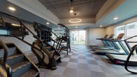 Located on Lankershim Boulevard in North Hollywood, this fitness center offers a comprehensive workout environment spread across a single level, seamlessly blending indoor and outdoor training spaces. The interior is designed with a modern aesthetic, featuring a checkered tile floor, recessed lighting, and a distinctive circular pendant light that anchors the cardio zone. Large floor-to-ceiling windows and sliding glass doors flood the space with natural light while providing direct views of and access to the outdoor turf area.

The cardio section is lined with treadmills, stair climbers, and elliptical machines positioned along the windowed walls, maximizing views and airflow. A dedicated strength training zone houses a multi-station cable machine, barbells, weight plates, and dumbbell racks, complemented by full-length mirrors that enhance the sense of space. A separate room with frosted glass partition walls offers a more private setting for stretching or focused training, fitted with a bench and additional equipment storage.

The outdoor turf area extends the training experience outside, equipped with plyo boxes, medicine balls, kettlebells, and a workout bench — ideal for functional and high-intensity training. The overall ambiance is clean, modern, and purposefully designed to support a wide range of fitness activities.