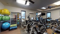 This fitness center is a thoughtfully designed, single-floor facility offering a thorough range of exercise equipment suited for a variety of workout routines. The space features durable cork and rubber flooring throughout, providing comfort and stability during training sessions. A two-tone wall treatment — warm white above and dark gray below — gives the room a clean, modern aesthetic.

The cardio zone is well-appointed with multiple Matrix treadmills, elliptical trainers, and a stationary bike, each positioned near windows that bring in natural light and exterior views. A wall-mounted flat-screen television accompanies the cardio area for entertainment during workouts. A ceiling fan and a wall-mounted mini-split air conditioning unit ensure a comfortable environment year-round.

The strength and functional training area includes a full dumbbell rack, an adjustable weight bench, a multi-function cable and pull-up tower, and a stair climber. A dedicated storage rack holds stability balls in multiple sizes, resistance bands, agility equipment, and additional accessories, supporting diverse training modalities. Mirror panels along one wall enhance the sense of space and allow users to monitor their form. The room is well-lit with overhead fluorescent fixtures, and access is provided through a keyed entry door with a sidelight window.