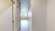 This studio in Seattle, WA offers a thoughtfully laid out single-floor plan that maximizes every square foot. The open-concept kitchen and living area serves as the heart of the space, featuring light wood-style flooring that runs throughout and large windows that invite natural light and frame views of the surrounding Seattle neighborhood. The kitchen is well-equipped with warm wood cabinetry, white countertops, and a full suite of stainless steel appliances including a refrigerator, electric range, built-in microwave, and dishwasher. Recessed track lighting illuminates both the kitchen and living areas effectively. A hallway connects the main living space to the rest of the unit, lined with two closets that provide generous storage for a studio layout. The bathroom features neutral tile flooring, a vanity with wood cabinetry, and a clean, functional design. Throughout the unit, the bright white walls and light flooring create a cohesive, airy aesthetic. The space is finished with floor-level heating vents and practical fixtures, making it a comfortable and efficient urban residence in Seattle's 98105 neighborhood.