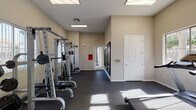 This fitness center is a dedicated, single-floor exercise facility designed to support a comprehensive workout routine. The space is finished with durable dark rubber flooring throughout, providing a safe and practical surface ideal for all types of training activities. Flush panel ceiling lights and large windows allow natural light to fill the room, creating a bright and energizing environment.

The cardio zone is equipped with a treadmill, elliptical trainer, and stationary bike, catering to a variety of aerobic exercise preferences. A wall-mounted flat-screen television is positioned for easy viewing during cardio sessions. The strength training area features a multi-functional cable machine and a Smith machine complete with a weight bench and free weights, offering users the tools needed for a full resistance workout.

The facility includes multiple entry and exit points, including a glass-paneled interior door that connects to an adjacent common area, as well as an exterior door providing direct outdoor access. Vertical blind-covered windows on multiple walls bring in natural light while maintaining privacy. The neutral beige walls and clean, minimalist aesthetic contribute to a focused and functional atmosphere throughout the space.