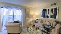 This single-floor residence offers a thoughtfully arranged layout that balances comfort and functionality across its living and sleeping areas. The floor plan reveals a clear separation between the main living space and the private bedroom, connected by a transitional corridor that also provides access to additional utility areas.

The living room serves as the heart of the unit, featuring soft neutral-toned walls, a textured ceiling, and carpet flooring that adds warmth underfoot. A large window dressed with vertical blinds allows generous natural light to fill the room, creating a bright and welcoming atmosphere. The space comfortably accommodates a sofa, accent chair, glass coffee table, and area rug, demonstrating its capacity for both relaxed lounging and casual entertaining. Wall art and decorative accents contribute to the room's cohesive, styled aesthetic.

The bedroom, visible in the floor plan overview, is a well-proportioned private retreat furnished with a full bed and flanking nightstands. Supporting spaces, including what appears to be a bathroom and closet or storage area, are accessible along the corridor, ensuring the layout remains practical and efficient throughout.