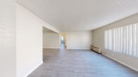 Located at 3149 E Desert Inn Rd in Las Vegas, NV, this single-floor unit offers a clean, functional layout with a bright and neutral aesthetic throughout. The open living area is finished with updated light gray wood-look flooring and smooth white baseboards, creating a fresh, modern feel. A textured popcorn ceiling adds dimension, while a wall-mounted HVAC unit ensures year-round comfort. Natural light filters in through windows dressed with vertical blinds, enhancing the airy atmosphere of the space. The living area flows seamlessly into a secondary room, providing flexible use of the layout. The kitchen is well-appointed with white cabinetry offering generous upper and lower storage, white appliances including a refrigerator, electric range, and dishwasher, and a peninsula with a white countertop that defines the space. A double-basin stainless steel sink sits beneath upper cabinets, and a ceiling fan in the adjoining dining nook promotes air circulation. An additional room features the same wood-look flooring and a window with views to the outside. Throughout, the consistent neutral color palette and updated finishes create a cohesive, move-in ready environment suited for everyday living in the heart of Las Vegas.