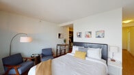 Located on the 44th floor of 345 E Wacker Drive in Chicago's vibrant Streeterville neighborhood, this well-appointed studio offers a thoughtfully designed open-plan layout that maximizes every inch of space. The main living area is anchored by a king-size bed dressed in crisp white linens and accented with warm mustard and gray tones, complemented by a sleek dark upholstered headboard. A cozy seating area with a mid-century inspired armchair and side table adds a relaxed lounge element to the space. A series of colorful modern art pieces adorns the white walls, lending the unit a vibrant, contemporary character. Rich hardwood-style flooring runs throughout, adding warmth and continuity. The compact kitchenette is outfitted with dark cabinetry and a full suite of stainless steel appliances, including a gas range, over-the-range microwave, and full-size refrigerator, alongside a small dining table ideal for casual meals. The bathroom features light tile flooring, a dark wood vanity with ample storage, a large mirror, and a glass-enclosed shower, offering a clean and functional design. The unit's efficient layout and high-floor positioning make it an ideal urban retreat in the heart of Chicago.