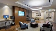 This single-floor space presents a polished, contemporary professional environment thoughtfully designed to balance functionality with refined aesthetics. The main lounge area serves as a central focal point, anchored by a bold circular pendant light fixture that adds a dramatic architectural element to the ceiling. Recessed lighting throughout complements this statement piece, ensuring the space is well-illuminated.

Rich wood wall paneling provides a warm backdrop to a wall-mounted flat-screen display, adding both visual depth and a sense of executive sophistication. The seating arrangement features structured blue upholstered armchairs paired with a dark, rounded coffee table, creating an inviting yet professional atmosphere. A neutral area rug defines the lounge zone against the herringbone-patterned hardwood flooring, which adds an elegant textural contrast.

A workstation is neatly integrated along one wall, offering a practical element without disrupting the refined aesthetic. Adjacent to the lounge, a glass-walled conference room is visible, offering transparency and an open feel while maintaining a distinct, dedicated meeting space with its own seating and artwork. Together, the design elements communicate a cohesive, upscale professional environment.