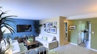 This single-floor residence offers a thoughtfully designed living space that balances color, comfort, and functionality. The living room immediately draws the eye with its striking deep blue accent wall, which contrasts beautifully against the neutral beige and sage green tones found throughout the space. A wall-mounted flat-screen television anchors one side of the room, while a curated gallery of framed artwork adds a personalized, artistic touch to an adjacent wall. The living area is furnished with a plush white sofa accented with decorative pillows, complemented by a dark wood coffee table and a bold geometric-patterned area rug in green and white tones. Lush indoor plants add a natural, refreshing element to the room. The layout flows openly toward a hallway that leads to additional rooms, including what appears to be a bedroom highlighted by a green accent wall and framed artwork. The textured popcorn ceiling and wall-to-wall carpeting are consistent throughout, contributing to a warm and cozy ambiance. Overall, the space presents a cheerful, well-coordinated interior with distinct character, making it both functional and visually engaging.
