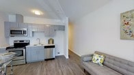 This studio unit with one bathroom is thoughtfully designed to maximize functionality within a compact footprint. The open-concept layout seamlessly integrates the living and kitchen areas, creating a cohesive and efficient living environment.

The kitchen is outfitted with sleek, high-gloss gray cabinetry that extends to the upper and lower walls, providing ample storage. Stainless steel appliances, including a dishwasher, complement the clean aesthetic, while a single-basin sink with a modern faucet adds to the unit's practicality. Light countertops contrast nicely with the gray cabinetry, enhancing the bright, airy feel of the space.

The living area flows naturally from the kitchen, featuring warm-toned hardwood-style flooring that runs throughout the unit, adding continuity and warmth. White walls and a white ceiling with flush-mount lighting keep the space feeling open and well-lit. Structural beams along the ceiling add a subtle architectural detail without compromising the clean lines of the interior.

The overall ambiance is modern and neutral, offering a versatile canvas well-suited for a range of interior styles. The efficient single-floor layout makes navigation straightforward, and the cohesive design elements throughout ensure a polished, move-in-ready feel.