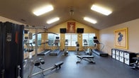 This single-floor fitness center offers a thoughtfully arranged workout space designed to support a variety of exercise routines. The room features a striking vaulted ceiling that adds a sense of openness and volume, complemented by warm yellow walls that create an energetic yet welcoming atmosphere. Overhead fluorescent lighting ensures the space is well-lit throughout.

The cardio area is well-stocked with multiple treadmills and elliptical machines, each equipped with individual display consoles. Strength training is supported by a cable and weight machine positioned along one wall, alongside a flat workout bench. Rubber flooring throughout the space provides both durability and safety during exercise.

Two flat-screen televisions are mounted on the walls, offering entertainment during workouts, while a wall clock keeps users on schedule. A decorative vintage-style poster adds a touch of character to the space. A large window along the back wall allows natural light to filter in, enhancing the overall ambiance. A wall-mounted mirror on one side visually expands the room and aids in form correction during strength exercises.