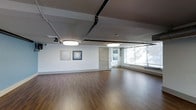 This single-floor studio space offers a clean, versatile layout suited for a variety of uses, from fitness and wellness activities to creative or professional gatherings. The room features warm hardwood flooring that extends throughout, providing a polished yet durable surface underfoot. White walls create a neutral backdrop, currently accented by two framed art pieces that add a subtle decorative touch without overwhelming the space.

The ceiling showcases an industrial aesthetic, with exposed black piping running overhead alongside two large circular pendant light fixtures that provide consistent, even illumination throughout the room. A projector is also mounted near the ceiling, suggesting the space is equipped for presentations or media use.

Large windows along one wall invite abundant natural light, creating a bright and airy atmosphere while offering views of the surrounding greenery. A standard interior door provides access to an adjacent area, contributing to the functional flow of the layout. The overall ambiance is minimalist and adaptable, combining clean design with practical infrastructure to accommodate a wide range of activities and configurations.