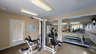 The fitness center at 1100 Autumn Creek Court in Manchester, Missouri, offers a well-equipped, single-floor exercise facility designed to support a range of workout routines. The space is organized across two connected areas, each serving a distinct fitness purpose.

The main strength training area features a multi-station cable machine with attached benches for leg and upper body exercises, along with instructional charts mounted on the warm-toned beige walls. Large wall mirrors expand the sense of space visually and allow users to monitor their form during workouts. Overhead fluorescent lighting provides consistent, even illumination throughout. A workout chart display and dumbbell storage are also present in this section.

The cardio zone houses two treadmills, a recumbent stationary bike, and an elliptical trainer, all positioned on durable carpet with protective floor mats beneath each machine. A wall-mounted television and a clock keep users informed and entertained during sessions. A decorative border runs along the lower portion of the walls, adding a subtle design element to the space.

Natural light enters through multiple windows fitted with plantation shutters, and views toward an outdoor pool area are visible from the interior. Multiple entry and exit doors provide convenient access, and the facility connects to an exterior staircase, indicating its role as a shared community amenity.