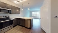 This single-floor residence offers a thoughtfully designed layout that maximizes functionality and comfort. The open-concept kitchen and living area serves as the heart of the home, featuring rich dark-toned cabinetry paired with light stone-inspired countertops and a peninsula that provides additional prep space and casual seating. A full suite of stainless steel appliances—including a refrigerator, range, microwave, and dishwasher—completes the kitchen. Pendant lighting and a track lighting system illuminate the space, while wide-plank hardwood-style flooring extends throughout the main living area. A large window in the living space allows natural light to flow in and offers views of the surrounding exterior.

The bedroom is a quiet, carpeted retreat with a dedicated window fitted with blinds, providing both natural light and privacy. Multiple doors suggest access to closet storage and connecting spaces. The bathroom is finished with dark tile flooring that complements the vanity cabinetry, which mirrors the kitchen's design. A granite-style countertop, undermount sink, large mirror, and a combination tub/shower complete the bathroom. Clean white walls and neutral tones throughout create a cohesive and move-in-ready environment.