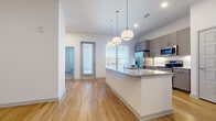 This thoughtfully designed two-bedroom, one-bathroom residence in Northlake, Texas offers a clean, contemporary aesthetic with quality finishes throughout. The open-concept living and kitchen area forms the heart of the home, where warm wood-look flooring flows seamlessly underfoot. The kitchen is a standout feature, equipped with a full suite of stainless steel appliances — including a French door refrigerator, range, microwave, and dishwasher — complemented by granite countertops, a central island with pendant lighting, gray shaker-style cabinetry, and a textured tile backsplash. Natural light pours in through multiple windows dressed with horizontal blinds, enhancing the bright, airy ambiance. The spacious primary bedroom features plush carpeting, a ceiling fan, and direct access to a generously sized walk-in closet outfitted with built-in shelving and hanging rods. The second bedroom also offers carpet flooring, a ceiling fan, and ample natural light. The full bathroom includes a soaking tub with tiled surround, while wood-look flooring extends into this space for a cohesive look. A dedicated living area with hardwood-style flooring and access to an outdoor balcony rounds out this well-appointed residence.