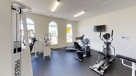 This fitness center is a thoughtfully designed, single-floor facility offering a comprehensive workout environment for users of all fitness levels. The space features crisp white walls, dark rubber flooring, and bright fluorescent lighting that creates a clean, energizing atmosphere throughout.

The facility is organized into distinct workout zones. The cardio area is equipped with treadmills and elliptical machines, complemented by a wall-mounted flat-screen television for entertainment during workouts. Large arched windows along one wall allow natural light to fill the space, creating an open and inviting feel.

The strength training area is well-stocked with a variety of professional-grade equipment, including Cybex and Precor cable and weight machines, as well as an adjustable bench and additional resistance training stations. The layout provides ample space to move between machines comfortably.

The facility also includes a dedicated stretch and functional training area, with equipment arranged to maximize usable floor space. Access to adjacent rooms is available through interior doors, suggesting additional amenities nearby. Ceiling-mounted speakers and climate control vents contribute to overall user comfort. The facility's clean, modern aesthetic and diverse equipment selection make it suitable for a wide range of fitness routines.