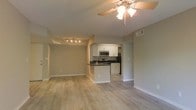 This single-floor space showcases an open-concept design that integrates the living area and kitchen into one cohesive, flowing layout. Light-toned hardwood flooring spans throughout, lending a clean and modern aesthetic to the interior. The living area is bright and airy, featuring neutral gray-beige walls and a ceiling fan with a multi-bulb light fixture that provides both comfort and illumination. A track lighting fixture adds a secondary layer of ambient light, enhancing the overall brightness of the space.

The kitchen is positioned adjacent to the living area, separated by a peninsula counter with a dark contrasting countertop that doubles as a breakfast bar. White cabinetry lines the kitchen walls, complemented by stainless steel appliances including a microwave, stove, and refrigerator. A sink is integrated into the peninsula, adding functional convenience. The kitchen benefits from its own recessed overhead lighting and features consistent hardwood flooring that ties it visually to the rest of the space. Neutral wall tones and clean white trim throughout contribute to a cohesive, well-maintained appearance.