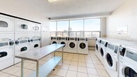 Located at 43 Forest Ave in Hamilton, Ontario, this shared laundry facility offers a clean, well-organized space designed for practical, everyday use. The room features light-coloured tile flooring throughout and bright white walls that contribute to a clean, functional aesthetic. Large floor-to-ceiling windows along one wall flood the space with natural light and provide sweeping elevated views of the Hamilton city skyline, making this a notably pleasant environment for a utility room.

The facility is equipped with a generous number of front-loading washing machines and stacked dryers arranged along the walls, offering ample capacity for building residents. A large stainless steel folding and sorting table is positioned centrally in the room, providing convenient workspace for laundry tasks. A second lower shelf beneath the table offers additional utility.

Safety and convenience features are well represented: a fire extinguisher is mounted on the wall, posted instruction signage assists users with machine operation, and a coin-operated or card-based payment terminal is available near the entrance. A seating bench along one wall provides a comfortable waiting area. The space is accessed through a solid utility door and is maintained in a tidy, accessible condition. Overall, this laundry facility is a functional and thoughtfully equipped communal amenity.