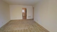 This single-floor space offers a straightforward and functional layout designed for comfortable everyday living. The main living area is generously sized, featuring wall-to-wall carpet in a warm beige tone that adds a soft, understated feel underfoot. The walls are finished in a clean, neutral cream palette throughout, creating a bright and cohesive atmosphere that lends itself well to a variety of décor styles. A floor vent provides climate control directly within the living space, ensuring year-round comfort. An open doorway connects the living area to an adjacent room, likely a kitchen or utility space, which benefits from natural light through a window visible in the background. This secondary area appears to include built-in appliances or cabinetry, suggesting a practical and efficient use of the space. The overall layout flows naturally from one area to the next, with clean sightlines and minimal structural obstructions. The design is simple and uncluttered, making it an adaptable space that can be easily personalized. The combination of neutral finishes, carpeted floors, and connected rooms creates a calm, livable environment suited for a range of occupants and furnishing arrangements.