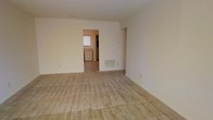 This single-floor space offers a straightforward and functional layout designed for comfortable everyday living. The main living area is generously sized, featuring wall-to-wall carpet in a warm beige tone that adds a soft, understated feel underfoot. The walls are finished in a clean, neutral cream palette throughout, creating a bright and cohesive atmosphere that lends itself well to a variety of décor styles. A floor vent provides climate control directly within the living space, ensuring year-round comfort. An open doorway connects the living area to an adjacent room, likely a kitchen or utility space, which benefits from natural light through a window visible in the background. This secondary area appears to include built-in appliances or cabinetry, suggesting a practical and efficient use of the space. The overall layout flows naturally from one area to the next, with clean sightlines and minimal structural obstructions. The design is simple and uncluttered, making it an adaptable space that can be easily personalized. The combination of neutral finishes, carpeted floors, and connected rooms creates a calm, livable environment suited for a range of occupants and furnishing arrangements.