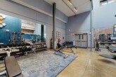This fitness center combines industrial architecture with modern gym functionality across a single, expansive floor. Soaring ceilings with exposed steel beams and structural columns create a dramatic vertical presence, while large upper-level windows allow natural light to filter into the space. The raw character of exposed brick walls — some painted in a deep teal blue — is complemented by polished concrete flooring and patterned area rugs that define distinct workout zones.

The strength training area is well-appointed with a comprehensive free weights section, featuring a full rack of dumbbells, adjustable benches, a multi-functional cable and smith machine, and dedicated mat space for functional training. Stability balls, barbells, and additional resistance equipment are neatly organized along the walls. Full-length mirrors line one side of the gym, visually expanding the space and providing feedback during workouts.

A separate cardio zone houses a row of modern treadmills, elliptical machines, stair climbers, and stationary bikes, all equipped with digital displays. Wall-mounted sconce lighting adds warmth throughout. An entry area with exposed brick, industrial-style wall fixtures, and double doors provides a distinct transition into the facility. The overall ambiance is energetic yet focused, blending the character of a converted industrial building with the functionality of a fully equipped modern gym.