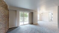 This single-floor space offers a thoughtfully arranged layout with a variety of functional and visually appealing features throughout. The open living and dining area is generously sized, finished with plush gray carpeting and neutral-toned walls that create a calm, versatile atmosphere. A sliding glass door floods the living area with natural light and provides direct access to a private outdoor balcony overlooking a tree-lined green space. A contemporary chandelier anchors the adjacent dining area, adding a stylish focal point to the open-plan layout.

The kitchen is well-equipped with two-tone cabinetry — white upper cabinets paired with gray lower cabinets — complemented by marble-style countertops, a stainless steel range with a sleek wall-mounted hood, and durable wood-look flooring. A separate area features a striking exposed brick accent wall, adding warmth and character to the space.

Bedrooms are carpeted and offer ample natural light through large windows, with at least one room featuring a sliding closet for convenient storage. The bathroom includes a tub-shower combination with subway tile surround, a white vanity with a single sink, a lighted mirror, and wood-look flooring, all brightened by a large window.