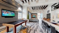 This contemporary single-floor clubhouse is thoughtfully designed to offer a sophisticated and welcoming community gathering space. The main lounge area commands attention with soaring ceilings adorned with bold dark exposed beams and an eye-catching array of globe pendant lights at varying heights, creating a dynamic yet refined atmosphere. Multi-panel arched windows flood the space with natural light, while wide-plank flooring in layered gray tones runs throughout, tying the open layout together cohesively.

At the heart of the entertainment area, a full-sized pool table rests atop a vibrant abstract area rug, anchored by a textured feature wall with a wall-mounted flat-screen TV. Nearby, a dedicated lounge zone offers plush sectional and accent seating arranged around striking hexagonal marble-top coffee tables, with an additional TV and warm wood credenza providing a comfortable media viewing area.

The kitchenette features dark cabinetry contrasted by crisp white countertops, a built-in microwave, and an open pass-through design with mirrored accents that visually expand the space. A separate dining area with a warm wood table and leather chairs provides an ideal setting for gatherings. Throughout the clubhouse, curated artwork, tall decorative planters, and carefully selected furnishings contribute to a polished, modern aesthetic.