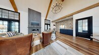 Located at 37 Foxgrove Dr in Dawsonville, GA, this single-floor clubhouse blends modern farmhouse aesthetics with sophisticated design across a generously sized open floor plan. The centerpiece of the main gathering area is a floor-to-ceiling gray wood-clad fireplace, flanked by black-framed French doors that invite abundant natural light and outdoor views. Dramatic vaulted ceilings with exposed rustic wood beams soar overhead, anchored by a striking geometric chandelier that adds an industrial-chic touch.

Rich hardwood flooring flows throughout the lounge and reception areas, transitioning to a bold black-and-white checkerboard tile in the kitchen and entry zones. A textured mosaic accent wall adds visual depth and character, complementing the navy blue cabinetry of the reception desk and bar area. The kitchen features a large white quartz island with seating, illuminated by pendant lighting with a warm, vintage feel.

Multiple seating arrangements — including tufted leather chairs, plush sofas, and intimate nook seating — offer flexible gathering spaces. Large black-framed windows throughout ensure the interior remains bright and connected to the surrounding community. Signage indicates access to fitness and restroom facilities, reinforcing the space's role as a full-service community amenity hub.
