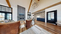 Located at 37 Foxgrove Dr in Dawsonville, GA, this single-floor clubhouse blends modern farmhouse aesthetics with sophisticated design across a generously sized open floor plan. The centerpiece of the main gathering area is a floor-to-ceiling gray wood-clad fireplace, flanked by black-framed French doors that invite abundant natural light and outdoor views. Dramatic vaulted ceilings with exposed rustic wood beams soar overhead, anchored by a striking geometric chandelier that adds an industrial-chic touch.

Rich hardwood flooring flows throughout the lounge and reception areas, transitioning to a bold black-and-white checkerboard tile in the kitchen and entry zones. A textured mosaic accent wall adds visual depth and character, complementing the navy blue cabinetry of the reception desk and bar area. The kitchen features a large white quartz island with seating, illuminated by pendant lighting with a warm, vintage feel.

Multiple seating arrangements — including tufted leather chairs, plush sofas, and intimate nook seating — offer flexible gathering spaces. Large black-framed windows throughout ensure the interior remains bright and connected to the surrounding community. Signage indicates access to fitness and restroom facilities, reinforcing the space's role as a full-service community amenity hub.