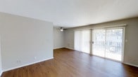 Located at 221 E. Main in Carmel, Indiana, this single-floor space offers a clean, straightforward layout designed for comfortable everyday living. The main living area is highlighted by warm hardwood-style flooring that runs throughout, adding a sense of warmth and continuity to the space. Neutral gray walls provide a versatile backdrop well-suited to a variety of décor styles.

A ceiling fan is centrally positioned overhead, contributing to both air circulation and ambient lighting. One of the most notable features of the living area is the full-width sliding glass door fitted with vertical blinds, which allows for generous natural light and direct access to an outdoor area, such as a patio or balcony. The outdoor view visible through the glass suggests a pleasant extension of the living space. A floor vent is also present, indicating a built-in climate control system. The overall ambiance is bright, open, and move-in ready, reflecting a simple and functional design well-suited to the Carmel, Indiana setting.