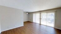 Located at 221 E. Main in Carmel, Indiana, this single-floor space offers a clean, straightforward layout designed for comfortable everyday living. The main living area is highlighted by warm hardwood-style flooring that runs throughout, adding a sense of warmth and continuity to the space. Neutral gray walls provide a versatile backdrop well-suited to a variety of décor styles.

A ceiling fan is centrally positioned overhead, contributing to both air circulation and ambient lighting. One of the most notable features of the living area is the full-width sliding glass door fitted with vertical blinds, which allows for generous natural light and direct access to an outdoor area, such as a patio or balcony. The outdoor view visible through the glass suggests a pleasant extension of the living space. A floor vent is also present, indicating a built-in climate control system. The overall ambiance is bright, open, and move-in ready, reflecting a simple and functional design well-suited to the Carmel, Indiana setting.