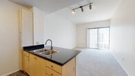 This single-floor residential unit offers a thoughtfully organized layout that blends functionality with comfortable living. The open-concept design connects the kitchen directly to the living area, creating a seamless flow throughout the main space. The kitchen features light wood cabinetry, dark granite-style countertops, hardwood-style flooring, a double-basin stainless steel sink, and black appliances including a full-size refrigerator and dishwasher. A peninsula counter extends into the living area, providing additional workspace and a natural visual divider between the two zones. The living area is finished with plush carpeting and is illuminated by track lighting overhead, with a sliding glass door fitted with vertical blinds that opens to an outdoor balcony, allowing natural light to fill the space. The bedrooms are carpeted and feature clean white walls, with windows that bring in additional natural light. The bathroom is well-appointed with an extended dark granite-top vanity, light wood cabinetry, dual mirrors, and a full-size soaking tub with a tiled shower surround and wood-style flooring. Throughout the unit, the neutral color palette and consistent finishes create a clean, cohesive ambiance.
