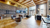 Located at 500 W 14th St in Richmond, Virginia, this single-floor communal space masterfully combines industrial heritage with modern comfort. The open-plan layout is anchored by soaring ceilings featuring richly toned exposed wood beams and planking, which lend a warm, loft-like character throughout. Original exposed brick walls serve as a defining architectural element, adding texture and depth to the space while honoring its historic roots.

Wide-plank hardwood floors stretch across the entire floor, unifying the various lounge areas beneath. Floor-to-ceiling glass doors and tall windows flood the interior with natural light and provide views of the surrounding neighborhood. A slatted wood partition wall, mounted with multiple flat-screen televisions, divides the space into distinct social zones without sacrificing openness.

Multiple seating arrangements accommodate a range of uses, from casual conversation to media viewing, featuring leather armchairs, upholstered sofas, and accent tables. A built-in bar counter with open shelving, pendant lighting, and bar stool seating offers a dedicated refreshment area. Statement chandeliers with gold accents complement the warm brass pendant lights, adding an elevated touch to the industrial aesthetic. The overall ambiance is both dynamic and welcoming, ideal for gathering and community engagement.