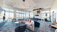 Located at 5455 Richmond Ave in Houston, TX, this contemporary community amenity space offers a sophisticated and well-appointed environment designed for social gathering and recreation. The single-floor layout encompasses a generously sized open-plan area that seamlessly integrates multiple functional zones.

The main lounge is a standout feature, anchored by a sweeping curved sectional sofa and complemented by an assortment of stylish accent chairs arranged around low-profile coffee tables. Overhead, dramatic starburst linear pendant chandeliers cast warm, ambient light across the soaring double-height ceilings, adding a striking architectural focal point. A floor-to-ceiling stone-clad fireplace with integrated display shelving serves as a commanding centerpiece along one wall.

A full-size billiards table occupies its own dedicated zone, offering a polished recreational option within the space. Adjacent to the lounge, a kitchenette area features rich navy cabinetry, a wet bar, and decorative tile backsplash beneath a bold patterned wallcovering.

Floor-to-ceiling windows and glass doors line the perimeter, flooding the interior with abundant natural light and framing panoramic views of the Houston city skyline. The geometric patterned carpet in deep blue tones ties the design together, lending cohesion to this elegantly curated amenity space.