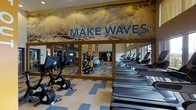 This single-floor fitness center is designed to energize and motivate members with a bold, ocean-themed aesthetic. The standout feature is a large, dramatic wall mural depicting crashing waves, accented by the bold "Make Waves" slogan rendered in prominent blue lettering — setting an active, dynamic tone throughout the space. The flooring features a striking blue and tan checkered pattern that adds visual interest and reinforces the aquatic theme. A row of modern treadmills lines one side of the space, positioned near windows that allow natural light to filter in, creating a bright and inviting workout environment. Stationary bikes and additional cardio equipment occupy the opposite side, offering a well-rounded selection of aerobic options. Full-length mirrors along the walls visually expand the space and allow exercisers to monitor their form. The mirrors also reflect additional strength-training equipment visible in the background, suggesting a more comprehensive equipment selection beyond the main floor. The overall ambiance is vibrant and purposeful, combining functional layout with motivational design elements to create an engaging environment suited to a wide range of fitness activities.