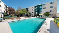 Located at 3901 Indian School Rd NE in Albuquerque, New Mexico, this outdoor amenity space is designed to maximize comfort and recreation under the renowned New Mexico sun. At the heart of the courtyard sits a generously sized swimming pool with crystal-clear turquoise water, surrounded by a spacious concrete sundeck lined with teal lounge chairs and colorful umbrella-shaded seating areas. The vibrant color palette — featuring teal, yellow, and blue accents — lends the space a lively, resort-like atmosphere. A centrally positioned gazebo provides a shaded retreat, complemented by additional hammock stations and casual seating arrangements ideal for relaxing outdoors. The landscaping incorporates a thoughtful mix of ornamental grasses, decorative gravel beds, mature trees, and manicured lawn areas, adding natural texture and greenery throughout. The courtyard is framed by a multi-story residential building with teal-accented balconies, creating an enclosed and private feel. A covered pergola with picnic-style seating offers another communal gathering area. Together, these features create a well-appointed, inviting outdoor environment that balances active recreation with peaceful relaxation.