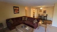 This single-floor space showcases an open-concept layout that seamlessly connects the living and dining areas, creating a cohesive and functional environment. The living area is anchored by a large, dark brown sectional sofa adorned with decorative pillows, paired with a patterned ottoman that doubles as a coffee table. Warm yellow-toned walls contribute to the cozy ambiance throughout the space, complemented by framed wall art that adds character. A ceiling fan with a light fixture is centrally positioned, providing both airflow and ambient lighting. Adjacent to the living area, a raised dining table with bar-style chairs offers a casual dining setting. Built-in shelving is visible near a hallway, providing additional storage and display options. The transition between the living and dining spaces flows naturally, making the area feel connected yet purposeful. The overall design emphasizes comfort and practicality, with neutral carpeting underfoot and a layout that maximizes the available space on the single floor.