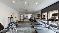 This fitness center offers a comprehensive workout environment spread across a single floor, designed to accommodate a wide range of exercise routines. The space is divided into distinct functional zones, each thoughtfully arranged to maximize usability and flow.

The cardio area is lined with Life Fitness treadmills and elliptical machines positioned along a bold, dark-accented wall with windows that bring in natural light and outdoor views. Wall-mounted televisions provide entertainment during workouts, and full-length mirrors amplify the sense of openness throughout the space.

A dedicated studio area features spin bikes, resistance training equipment, TRX suspension systems, kettlebells, and balance tools such as a BOSU ball, all arranged on a smooth hardwood-style floor. Motivational fitness artwork adorns the walls, and ceiling fans ensure comfortable air circulation throughout both zones.

The strength training section includes a multi-station cable machine, selectorized weight machines, and free weights, catering to a full-body workout experience. Patterned carpet tiles ground the equipment areas and add visual texture to the overall design.

Convenience amenities include dual stainless steel drinking fountains and clearly marked restroom facilities. Recessed lighting throughout provides bright, consistent illumination, creating an energetic and welcoming atmosphere for users of all fitness levels.