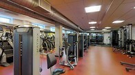 This fitness center offers a comprehensive workout environment designed to accommodate a wide range of exercise needs. The space features an industrial aesthetic, highlighted by exposed metal ductwork running along the ceiling, which adds a distinctive character to the room. Recessed and flush-mounted ceiling lights provide consistent, even illumination throughout.

The strength training area is well-stocked with a row of Matrix cable and resistance machines, each positioned to allow ample space for safe and comfortable use. Structural columns punctuate the floor plan, helping to naturally segment the space into distinct workout zones. Along one side, a selection of cardio equipment — including ellipticals and step machines — lines the area near windows that allow natural light to filter in.

The flooring consists of a durable red rubberized surface, ideal for high-traffic fitness use and providing cushioning underfoot. Large mirrors along the walls visually expand the space and allow users to monitor their form. The overall layout is organized and spacious, creating a functional and motivating environment for both strength and cardiovascular training.