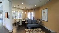 This thoughtfully designed single-floor space combines comfort and functionality in a warm, inviting atmosphere. The main area is anchored by a cozy lounge section featuring a plush gray sofa and accent seating, arranged atop a bold patterned area rug that adds visual interest to the rich, dark hardwood flooring beneath. Warm amber-toned walls lend a welcoming ambiance throughout, complemented by crisp white crown molding that frames the space with a polished finish. Adjacent to the lounge, a bar-height counter offers additional seating with upholstered stools, making it ideal for casual gatherings or collaborative use. Three elegant pendant lights hang above the counter, providing focused illumination and a touch of refined style. Natural light flows generously through multiple windows dressed with neutral blinds and draped curtains, creating a bright and airy feel. Recessed ceiling lights supplement the natural light, ensuring the space remains well-lit at any time of day. A framed art piece on the wall adds a creative focal point, while the mix of industrial-style metal chairs and softer furnishings strikes a balanced, contemporary aesthetic. Overall, the space is designed to be both functional and inviting for a variety of uses.