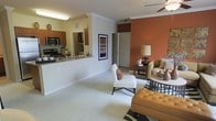 This single-floor residence showcases an open-concept design that integrates the living and kitchen areas into one cohesive, flowing space. The living area is anchored by a bold terracotta accent wall, which adds warmth and visual depth to the room. Plush carpeting runs throughout, contributing to a comfortable and inviting atmosphere, while a ceiling fan overhead ensures year-round comfort.

The kitchen is accessible via a raised breakfast bar with granite countertops, offering a practical casual dining option while maintaining visual connection to the living space. Warm wood cabinetry, a stainless steel microwave, and a gas range are among the notable kitchen features, complemented by tile flooring that contrasts with the carpeted living area.

Crown molding along the ceiling adds an elegant, finished touch throughout the space. A framed art piece on a neutral wall bridges the kitchen and living zones, enhancing the curated aesthetic. A closet door is visible toward the rear of the living space, hinting at thoughtful storage solutions. Overall, the residence balances warm, earthy tones with practical design for comfortable everyday living.