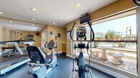 This single-floor fitness center offers a comprehensive workout environment designed to support a variety of exercise routines. The space is laid out in a rectangular floor plan with warm beige walls, durable rubber flooring, and a textured ceiling fitted with recessed lighting that keeps the room well-illuminated throughout. Large windows along one side of the room flood the interior with natural light and provide direct views of an outdoor pool and patio area, creating an inviting atmosphere for exercise. The cardio section is equipped with multiple machines, including a Spirit treadmill, an elliptical trainer, a stair climber, and a recumbent stationary bike, each positioned to take advantage of the pool-facing windows. A wall-mounted flat-screen television is available for entertainment during workouts. On the opposite end of the room, a multi-function cable weight machine with a bench and weight stack offers a dedicated space for strength training. A medicine ball and additional accessories are also on hand. Ceiling-mounted fans provide additional air circulation, and sanitation supplies are conveniently stationed near the entrance. Posted notices and signage indicate facility rules and guidelines. The overall ambiance is clean, functional, and purposefully designed to accommodate a well-rounded fitness routine.