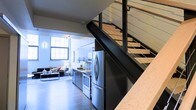 This two-story space showcases a thoughtfully designed open-concept layout that seamlessly blends modern and industrial aesthetics. The main level integrates the kitchen and living area, creating a cohesive, functional living environment. The kitchen is outfitted with stainless steel appliances, including a French door refrigerator and a range, complemented by streamlined cabinetry in a light finish. A galley-style counter configuration maximizes efficiency while maintaining visual openness.

The living area benefits from a large, multi-pane industrial-style window that draws in generous natural light, enhancing the bright, airy atmosphere of the space. Hardwood flooring runs throughout the main level, adding warmth to the otherwise contemporary palette of whites and dark accents.

A striking floating staircase serves as a central architectural feature, constructed with dark metal stringers and cable railing, lending the space a refined, urban character. The open-riser design allows light to pass through, maintaining visual connectivity between floors. The staircase leads to the upper level, expanding the overall functionality of the space. The combination of industrial detailing and modern finishes creates a dynamic, well-balanced environment throughout.