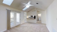This single-floor residence showcases an open and airy design highlighted by soaring vaulted ceilings that lend a sense of generous vertical space to the main living area. A ceiling fan with a built-in light fixture is centrally positioned overhead, complementing a skylight that draws in abundant natural light. Soft, neutral carpeting runs throughout the living space, creating a warm and inviting atmosphere. A pass-through kitchen counter connects the kitchen to the living area, offering a semi-open layout that encourages interaction between spaces. The kitchen is equipped with dark countertops, white cabinetry, and black appliances, including a range and microwave. A chandelier illuminates the dining area, which is visible just beyond the kitchen. French doors open to an outdoor deck, providing a seamless transition to the exterior and allowing natural light to pour into the room. Views of lush greenery are visible through the glass panes. A dedicated laundry area, complete with a washer and dryer, is conveniently located just off the main living space. The overall ambiance is clean, bright, and functional, with a neutral color palette throughout that creates a versatile and welcoming environment.