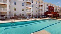 Located at 1000 Sweetbay Haven Ln in Apex, NC, this outdoor amenity space is thoughtfully designed to offer residents a resort-style living experience. The centerpiece of the area is a generously sized swimming pool with crystal-clear water, surrounded by a broad concrete sundeck furnished with lounge chairs and shaded umbrella seating — ideal for relaxing on sunny North Carolina afternoons.

The pool area is enclosed by a black iron fence, providing both security and an open, airy feel. Mature trees and young landscaping line the perimeter, adding natural greenery to the setting. The multi-story residential building frames the courtyard with a classic exterior featuring gray horizontal siding, white balconies, and warm wood-toned accents under the eaves, creating an inviting architectural backdrop.

A covered walkway runs alongside the building, equipped with ceiling fans that provide comfort during warmer months. Warm-toned wood entry doors with frosted glass sidelights open to individual units, while the corridor looks out toward the pool and green spaces. A lawn area is visible nearby, adding additional outdoor recreational space. The overall ambiance is bright, clean, and welcoming, blending functional amenities with attractive design elements suited to an active residential community.