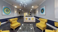 This well-appointed communal lounge space is thoughtfully designed with a cohesive navy blue and gray color palette that runs throughout. A distinctive two-tone wall treatment—navy on the lower half and soft gray above a crisp white chair rail—anchors the interior's sophisticated aesthetic, complemented by white crown molding along the ceiling line.

The main lounge area is generously proportioned, offering multiple seating arrangements centered around a striking navy built-in entertainment wall. This architectural focal point features a large flat-screen television framed within an arched millwork surround with detailed paneling. Bold yellow armchairs and a white upholstered sofa create vibrant contrast against the darker tones, while a geometric glass coffee table adds a modern touch. Pendant lights and a multi-arm chandelier provide layered illumination throughout.

A separate, more intimate seating nook offers a quieter retreat, furnished with contemporary lounge chairs arranged around a round marble side table. Abstract circular artwork adorns the walls of both areas, reinforcing a curated, gallery-like atmosphere. Herringbone-patterned carpet tiles in charcoal tones unify the entire space underfoot, contributing to its polished, professional character.