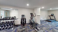 This fitness room is a thoughtfully designed, single-level space dedicated to supporting a well-rounded workout routine. The room is finished with neutral gray walls, white baseboards, and a geometric patterned carpet in shades of gray and blue, lending the space a clean, modern aesthetic. Recessed lighting illuminates the room evenly throughout.

The cardio area houses two treadmills positioned near a window that allows natural light and outdoor views, creating an energizing environment for users. Alongside the treadmills, a rowing machine and an elliptical trainer provide a range of low-impact cardio options.

A dedicated strength training area features a dumbbell rack stocked with free weights and an adjustable workout bench, suitable for a variety of resistance exercises. A large wall-mounted mirror in this zone enhances the sense of space and allows users to monitor their form. A TRX suspension training system is anchored to the wall, offering versatile bodyweight training options.

The room also includes a built-in shelf for personal items, a water cooler station with a trash receptacle, and a wall-mounted unit for climate control. A glass-panel door with a teal frame opens directly to an outdoor area, providing convenient ventilation and easy access.