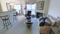 This single-floor residence offers a warm and inviting living area designed for both comfort and functionality. The living room is a central highlight, featuring a white-surround fireplace that serves as a natural focal point. Large windows dressed with decorative valances and horizontal blinds allow abundant natural light to fill the space, creating an airy atmosphere throughout.

The living room is furnished with a plush sofa adorned with patterned throw pillows, a leather recliner with matching ottoman, and a round glass-topped coffee table with a woven base, adding a touch of texture to the room. Lush indoor plants near the fireplace contribute a natural, organic element to the overall aesthetic. The flooring is carpeted in a neutral tone, enhancing the room's cozy feel.

Adjacent to the living area, a raised breakfast bar with tall barstools provides a casual dining option, seamlessly connecting the living space to what appears to be a kitchen area. Wicker accent chairs are also visible, reinforcing a relaxed, nature-inspired design theme. The overall ambiance is light, comfortable, and thoughtfully arranged.