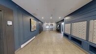 This building lobby presents a thoughtfully designed entrance space that balances functionality with contemporary style. The interior is defined by striking deep blue wainscoting and panel molding that lines the walls, contrasting cleanly against light grey upper walls and a white ceiling fitted with recessed pot lighting throughout.

Large-format neutral ceramic floor tiles extend across the full area, lending a sense of openness and ease of movement. The entrance features full-height glass doors with aluminum framing, allowing natural light to filter into the space and providing a clear sightline to the exterior.

A prominent built-in mailroom wall on one side houses an extensive array of individual resident mailboxes, finished in coordinating blue cabinetry with open lower shelving below. On the opposite end, two stainless steel elevator doors are flanked by white paneled walls, with a decorative accent vignette between them — featuring a round gold-framed mirror and a slim console table with decorative accessories — adding a refined residential touch.

The lobby also includes an intercom panel near the entrance and safety equipment discreetly integrated into the walls. Overall, the space conveys a clean, modern aesthetic with upscale finishing details.