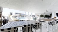 This sophisticated poolside space offers a seamless blend of covered comfort and open-air resort living. The venue is anchored by a sleek covered terrace with a smooth white ceiling, providing shade while maintaining an airy, open ambiance. Beneath the canopy, high-top dining tables crafted from slatted wood are paired with bold black-and-white striped chairs, creating a sharp, contemporary aesthetic that carries throughout the space.

The area opens directly onto a sprawling resort-style pool, which serves as the visual centerpiece. Rows of white sun loungers line the pool deck, while white fabric cabanas and umbrellas are arranged along the perimeter, offering guests additional shaded retreats. Lush potted tropical plants add natural color and warmth against the predominantly white palette.

The surrounding architecture features clean-lined, modern white buildings with large windows, reinforcing the resort's upscale, minimalist character. Light-colored tile flooring runs throughout, enhancing the bright and polished atmosphere. A ceiling fan is discreetly integrated into the covered section, ensuring comfort during warmer hours.

Overall, this space communicates a refined, resort-quality experience, ideal for dining and socializing with direct access to expansive pool and outdoor amenities.